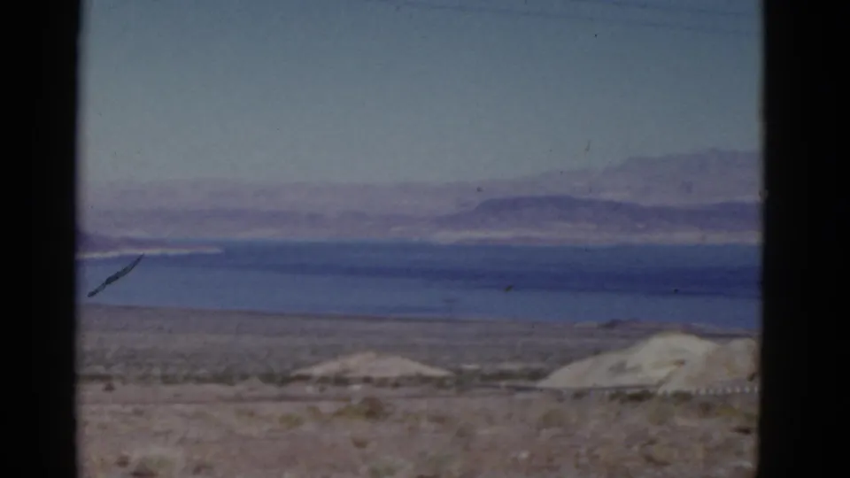 Faded 8mm California travel frame with scrubby desert foreground, pale mounds, water, and soft mountain shapes in the distance.