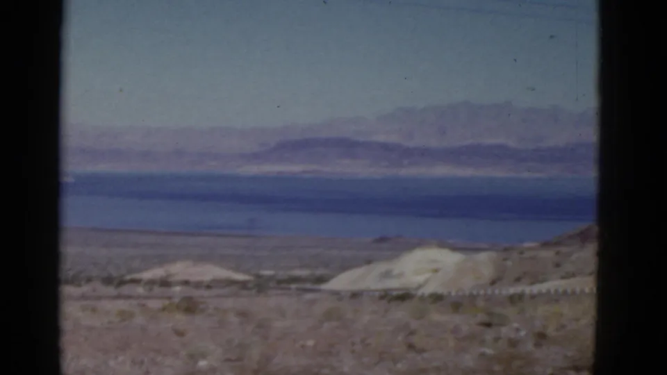 1952 California 8mm frame showing roadside desert terrain, a faint utility line, blue water, and mountains fading into haze.