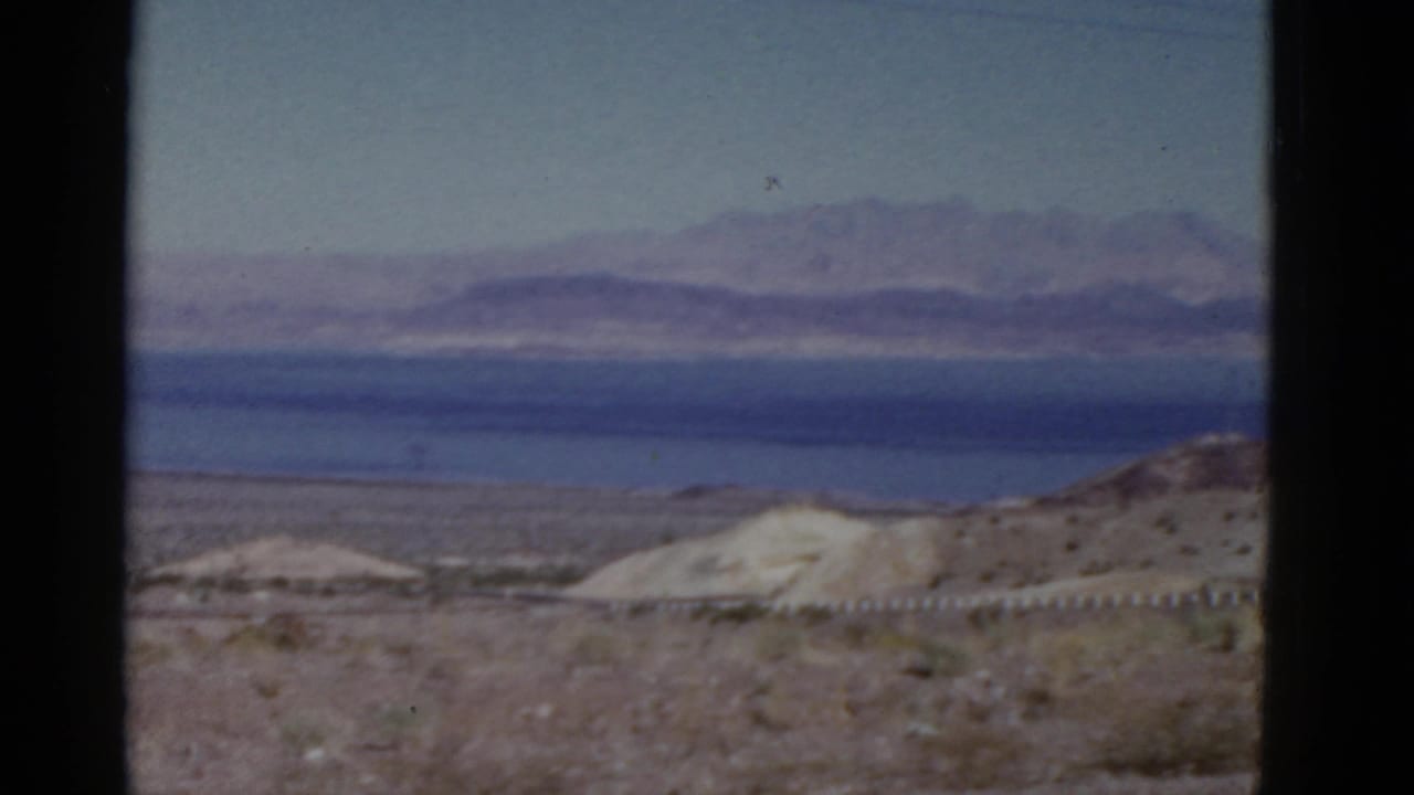 1952 California home-movie frame showing a broad desert view with pale hills, a distant blue basin or salt flat, and purple mountain ridges.