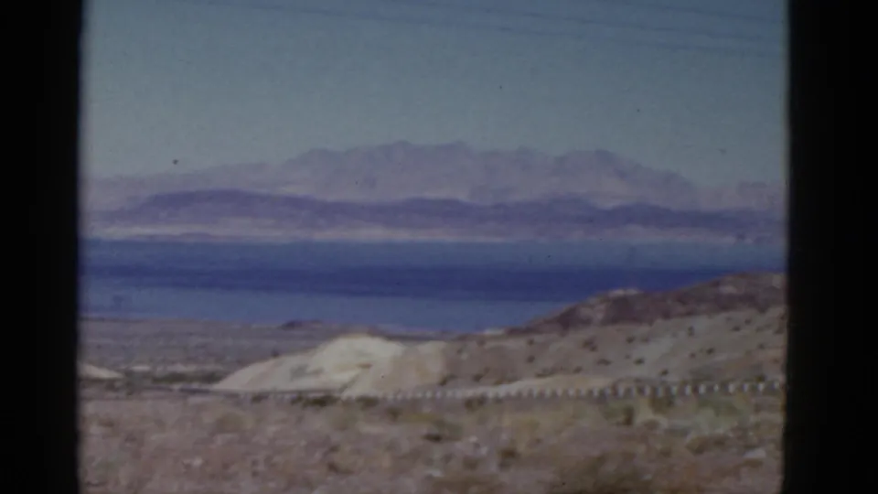 1952 California 8mm frame of arid roadside terrain looking toward pale hills, a blue distant expanse, and hazy purple mountains.