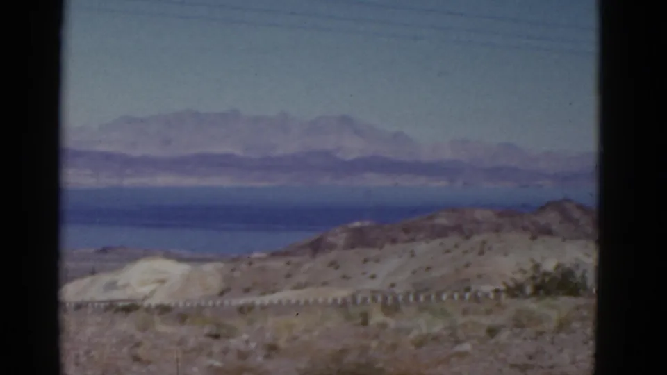 1952 California travel footage frame with scrubby desert ground, low pale hills, distant blue water-like or salt-flat area, and mountain ridges.