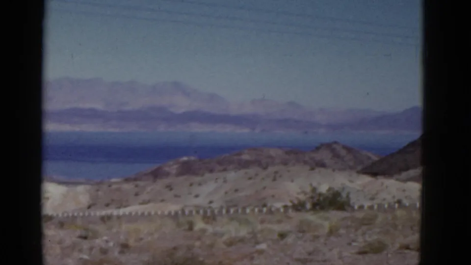 1952 California home-movie frame showing roadside desert brush and low hills against a blue distant expanse and hazy mountain backdrop.