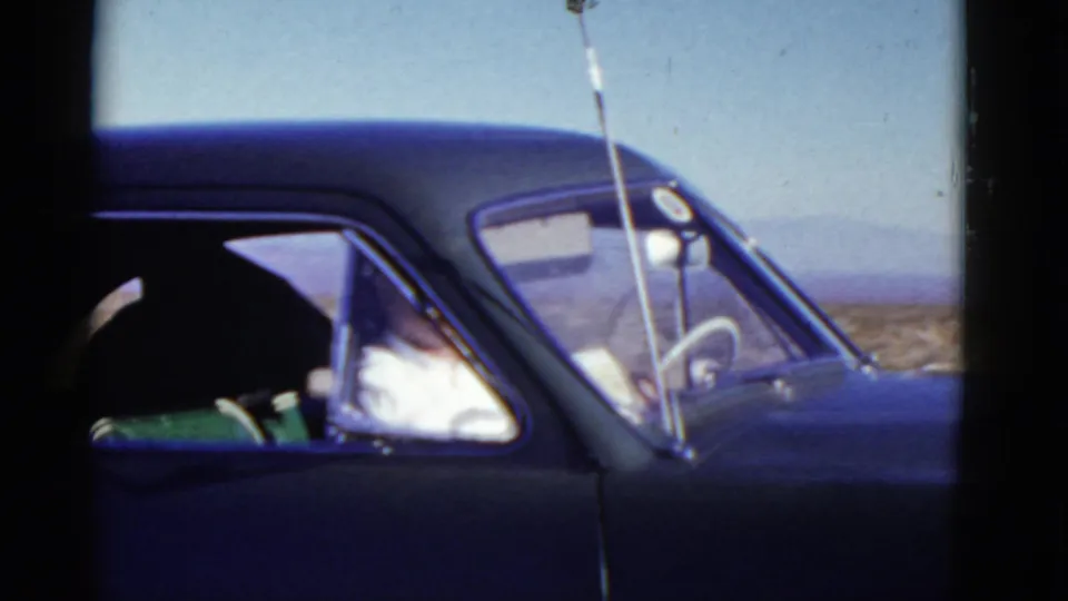 A dark mid-century car passes through the Death Valley approach, with an occupant visible through the side window.
