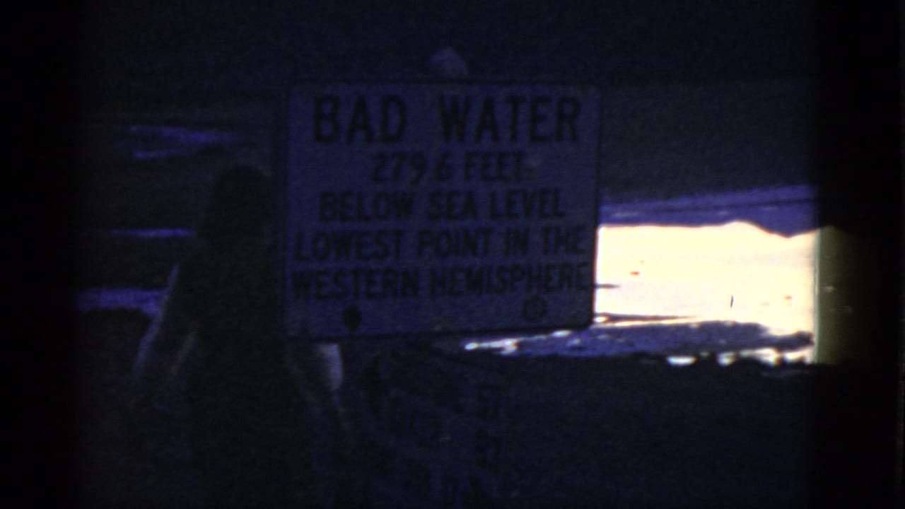 1952 California 8mm frame showing a silhouetted visitor near a Bad Water sign, with bright water and dark shoreline beyond.