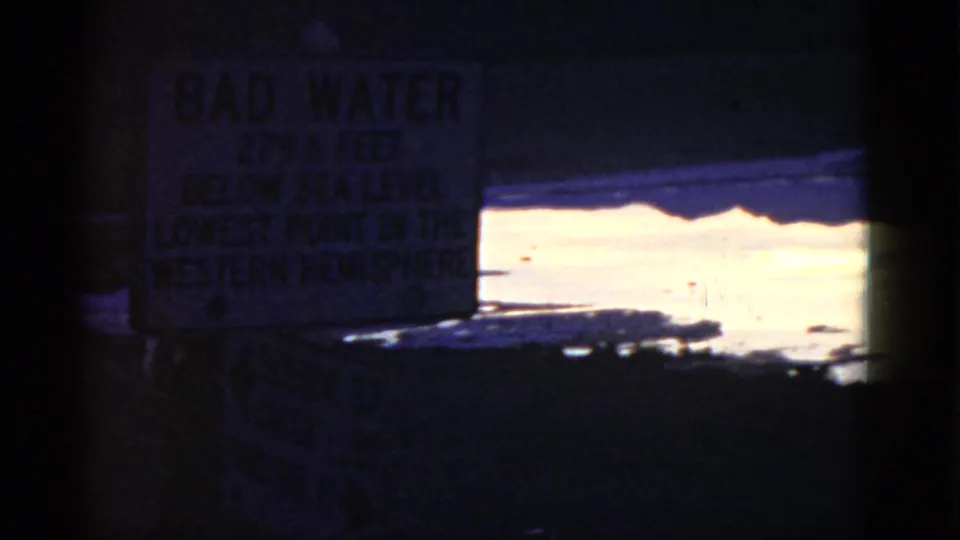 Bad Water sign in a dark 1952 California travel frame, with bright water, shoreline, and heavy film-edge shadowing visible.