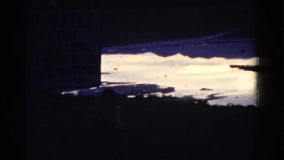 A cropped 8mm view of the Bad Water sign at left, with bright water, dark shoreline, and blue-tinted shadows in California.