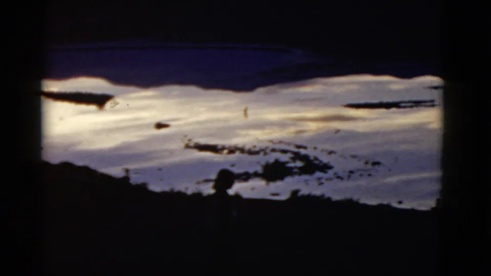 The vintage 8mm frame shows a bright snowy California slope with dark land patches and a distant silhouetted person at the lower right.