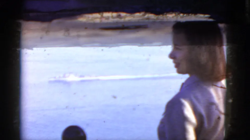 Young woman in profile watches the ocean as a distant ship or wake crosses the water in this 1952 California home-movie frame.