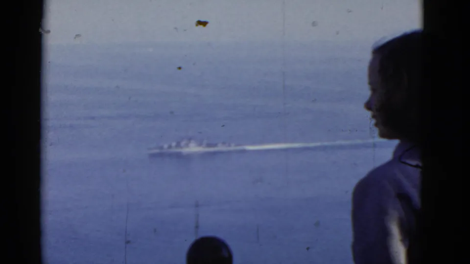 Silhouetted young woman looks toward the ocean while a distant vessel wake moves across the frame, with another person partly visible below.