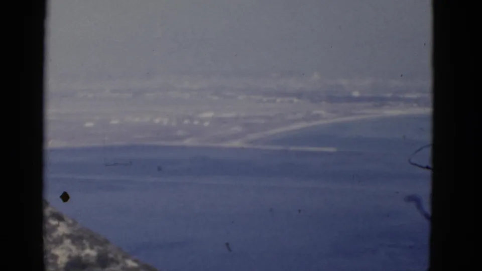 1952 California 8mm view of a hazy shoreline and distant buildings, with blue water and a rocky edge entering the frame.