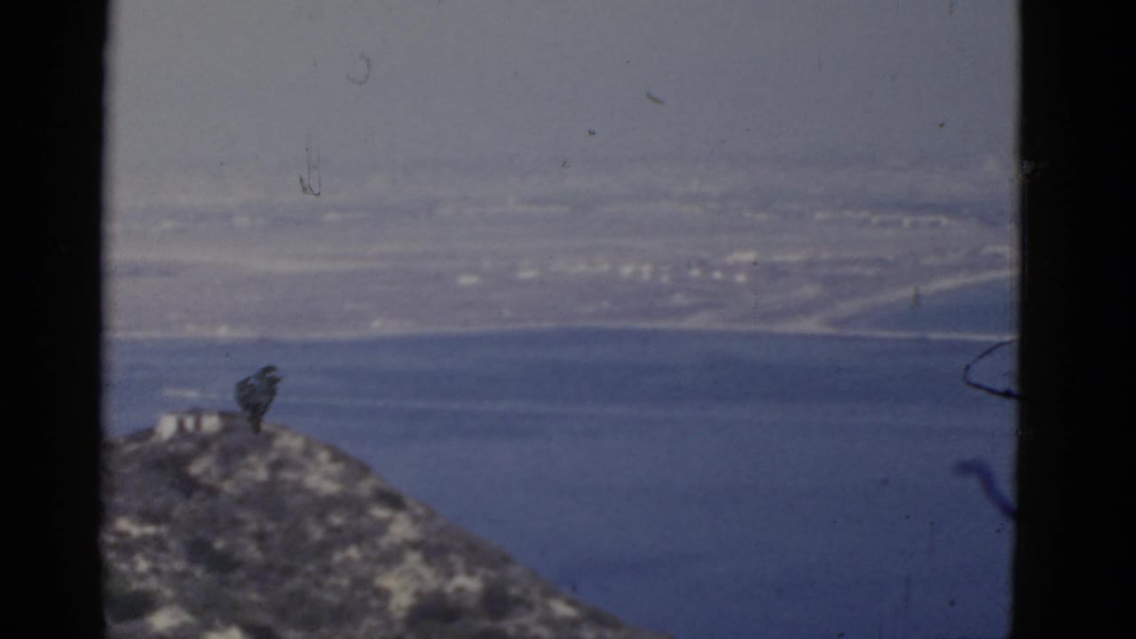 Rocky hillside above blue bay water with a hazy distant shoreline.