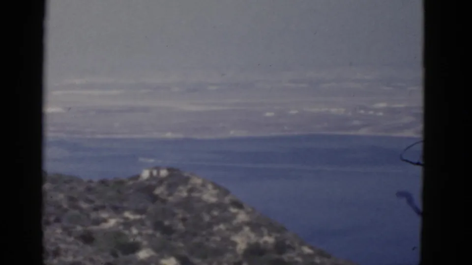 1952 California 8mm frame with a small light-colored hilltop structure, rocky slope, bay water, and hazy shoreline.