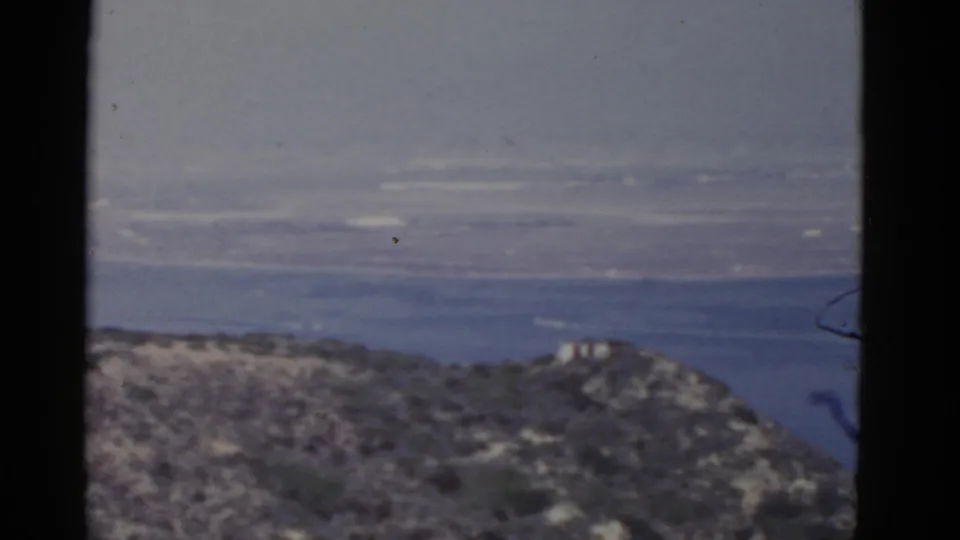 1952 California 8mm frame with rocky hillside foreground, a small distant structure, bay water, and hazy land beyond.