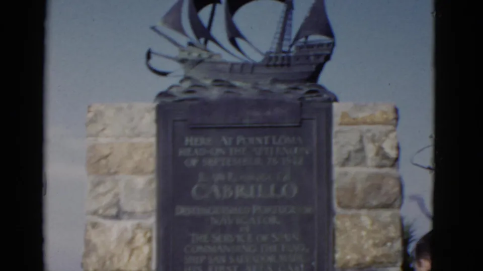 Cabrillo monument at Point Loma with plaque, ship sculpture, stone wall, and a partial visitor entering the edge of frame.