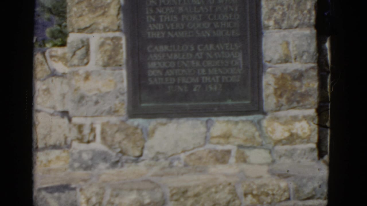 1952 California home-movie frame showing a dark historical marker set into a rough stone wall.