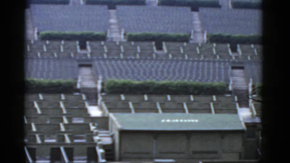 Mid-century 8mm frame showing a grid of gray bungalow housing rows with hedges and a green-roofed foreground building.