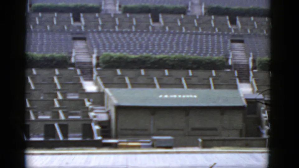 1952 California home-movie frame with rows of gray housing, hedge lines, concrete paths, and a green-roofed foreground structure.