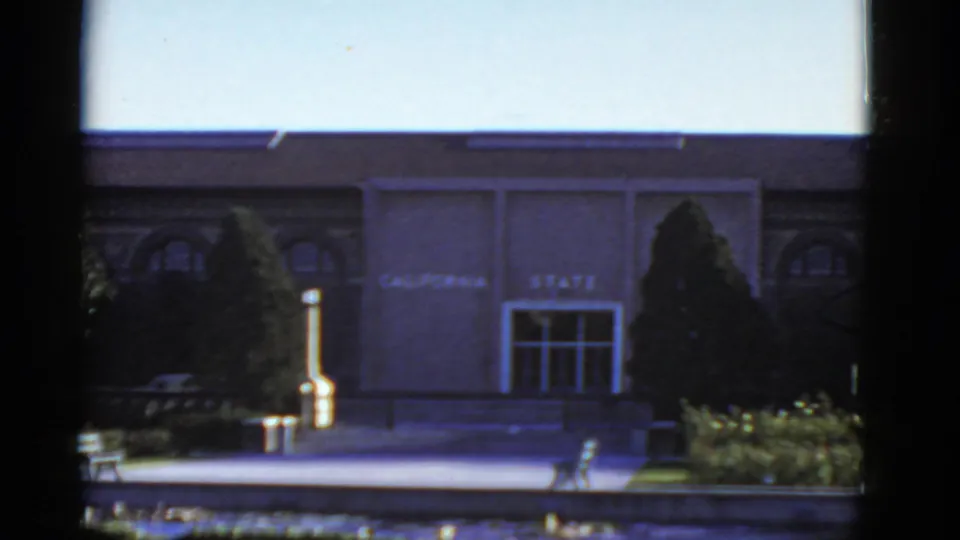 1952 California 8mm frame of a distant institutional entrance labeled "CALIFORNIA STATE," bordered by trees and low landscaping.