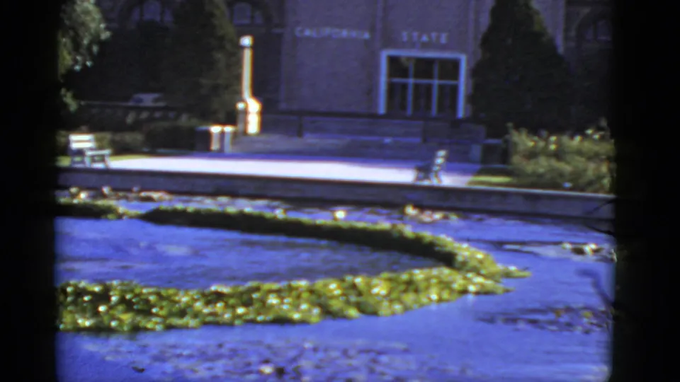 Home-movie frame of a California civic facade with entrance area, bench, lamppost, pond, lily pads, and manicured planting.