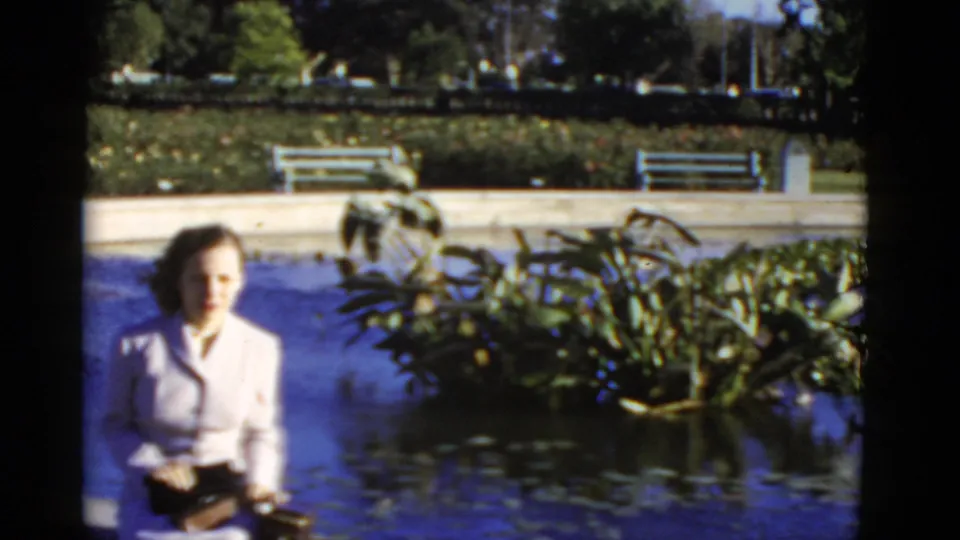 1952 California 8mm frame showing a woman in a light suit near a landscaped pond with plants, benches, and trees.