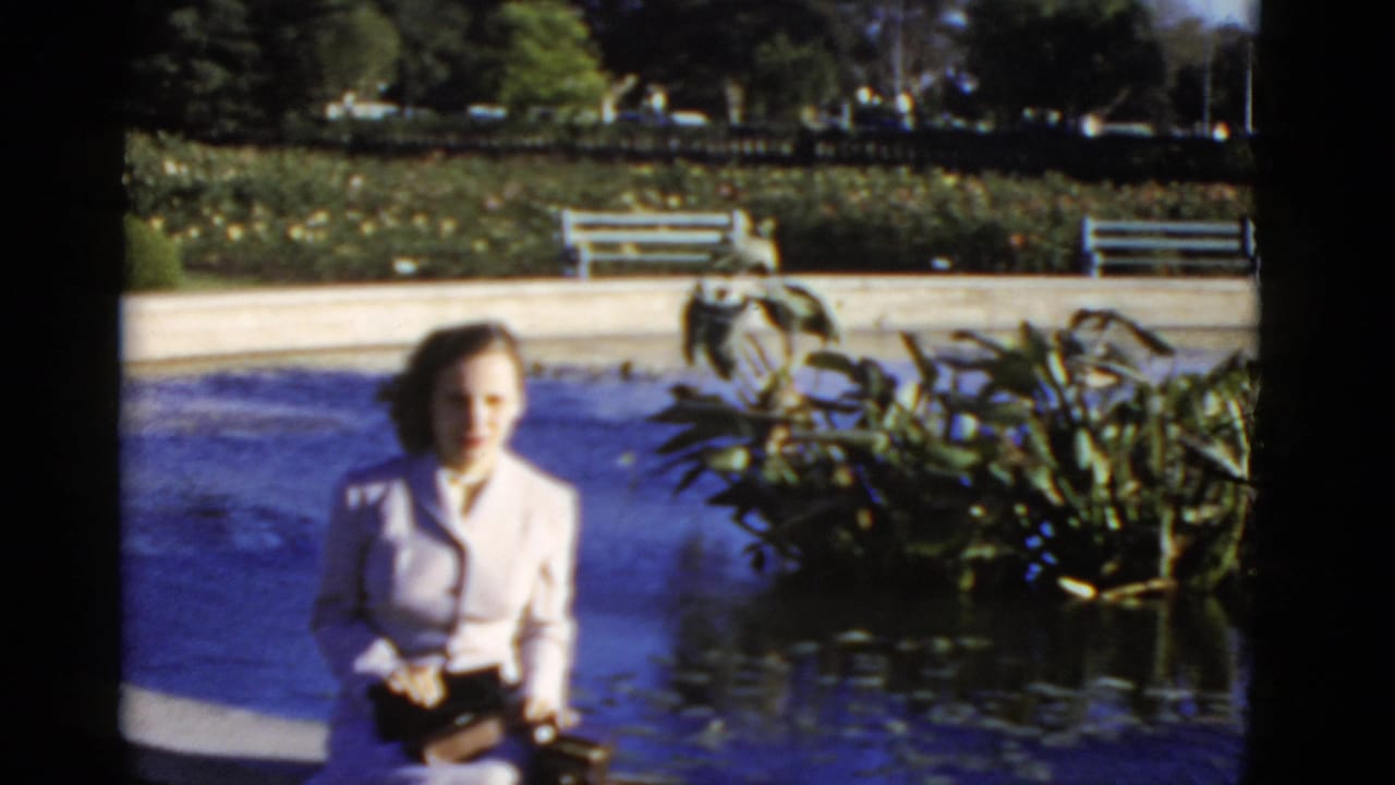 Woman in a light suit seated beside a circular pool with plants and benches behind her.