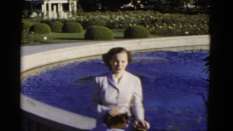1952 California home-movie frame showing a woman by a curved pool edge, with clipped hedges, flowers, and a white pergola beyond.