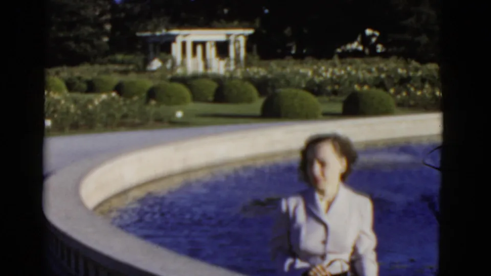Soft-focused 1952 California 8mm frame of a seated woman partly low in frame beside a circular pool and formal garden beds.