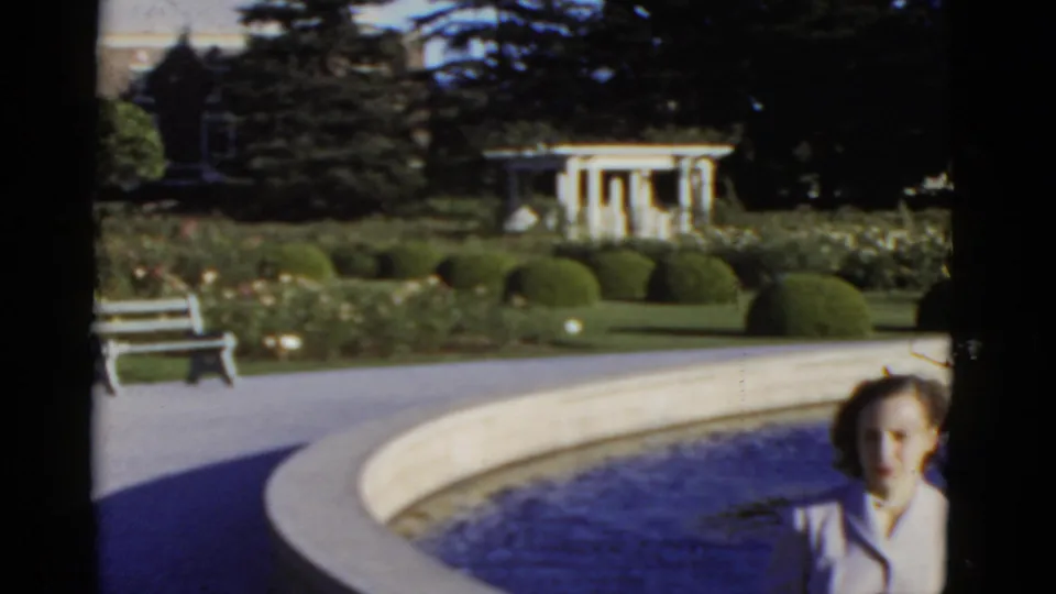 Wide 1952 California garden frame with a circular blue pool, white bench, clipped shrubs, pergola, and the seated woman near the lower right edge.