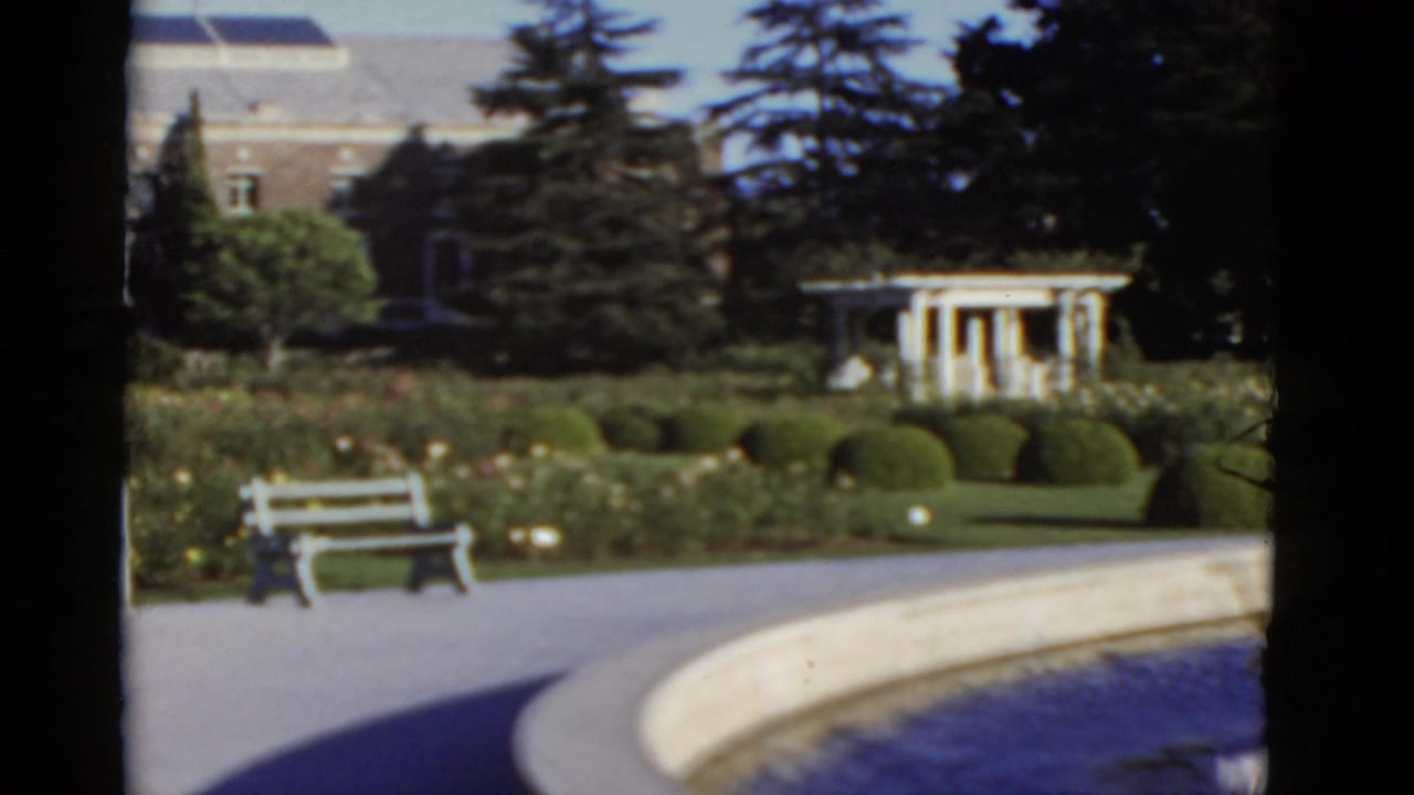 Courtyard with a white bench, curved fountain edge, hedges, trees, and a large building beyond.