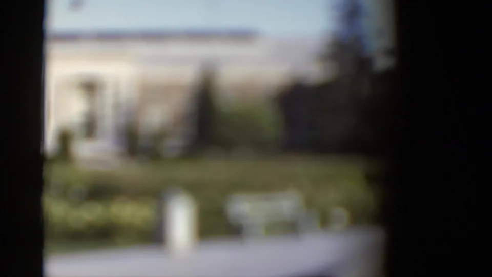 Soft-focus 1952 California frame with a pale building, garden hedges, a bench or low object, and dark vertical film edges.