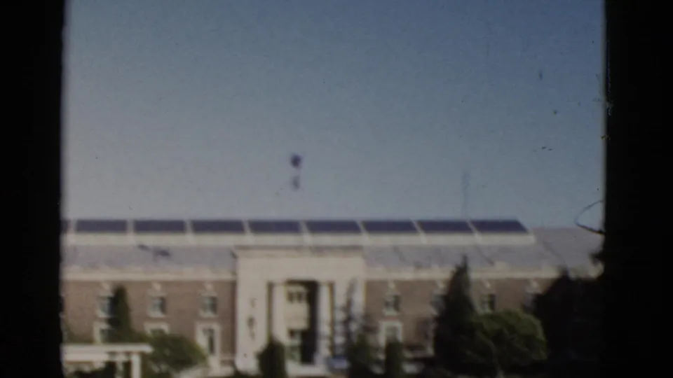 1952 California 8mm frame showing a large masonry building, columned entrance, dark roofline elements, trees, shrubs, and open sky.