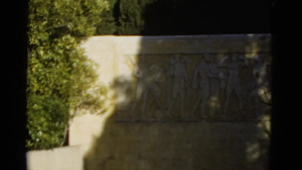 1952 California 8mm frame showing a sunlit stone memorial relief with athletic figures, foliage at left, and dark film edges.