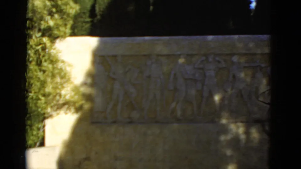 1952 California home-movie frame of a carved athletic relief on a pale monument wall, with trees and dappled shade across the surface.