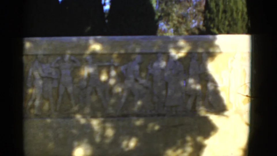Outdoor 8mm view of a stone frieze in California, with ancient-style figures, soft focus, tree shade, and a sunlit right side.