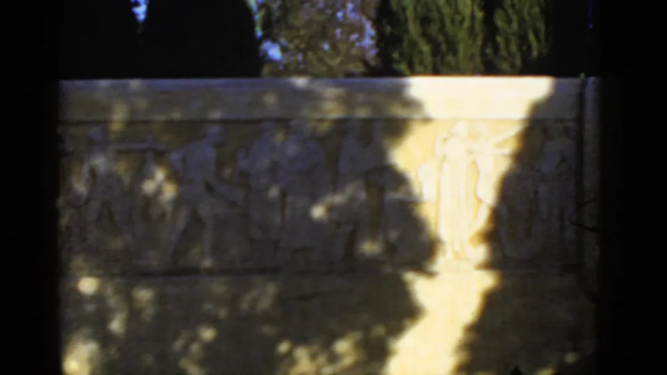 1952 8mm frame of a stone relief panel, with carved human-like figures obscured by shade and a bright sunlit strip.