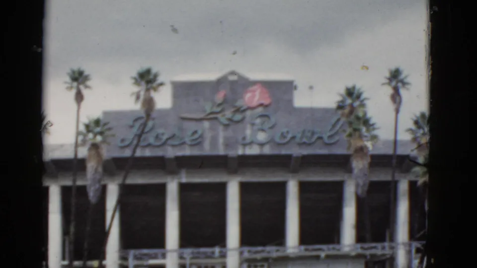 California home-movie frame of the Rose Bowl sign across a building front, with tall palms and cloudy sky behind it.