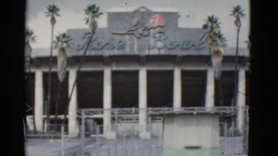 Handheld 8mm view of the Rose Bowl facade in California, with the sign, rose emblem, palms, and open columned front visible.