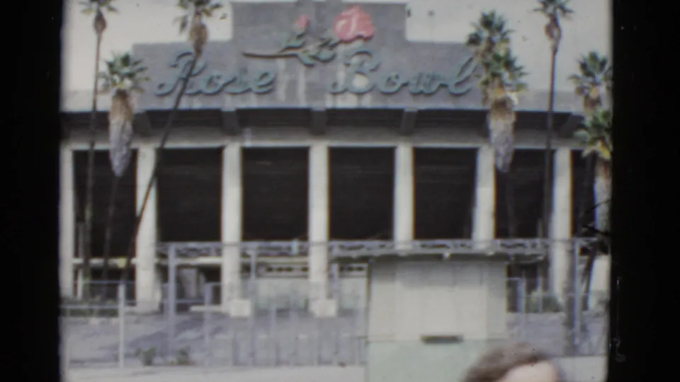 1950s 8mm view of the Rose Bowl exterior, showing the cursive sign above columns, palm trees, railings, and entrance steps.