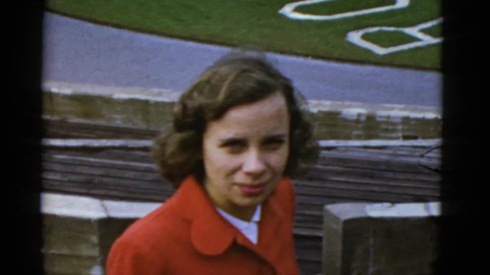 Young woman in a red coat smiles near the Rose Bowl grounds, with steps, pavement, green lawn, and white edging behind her.
