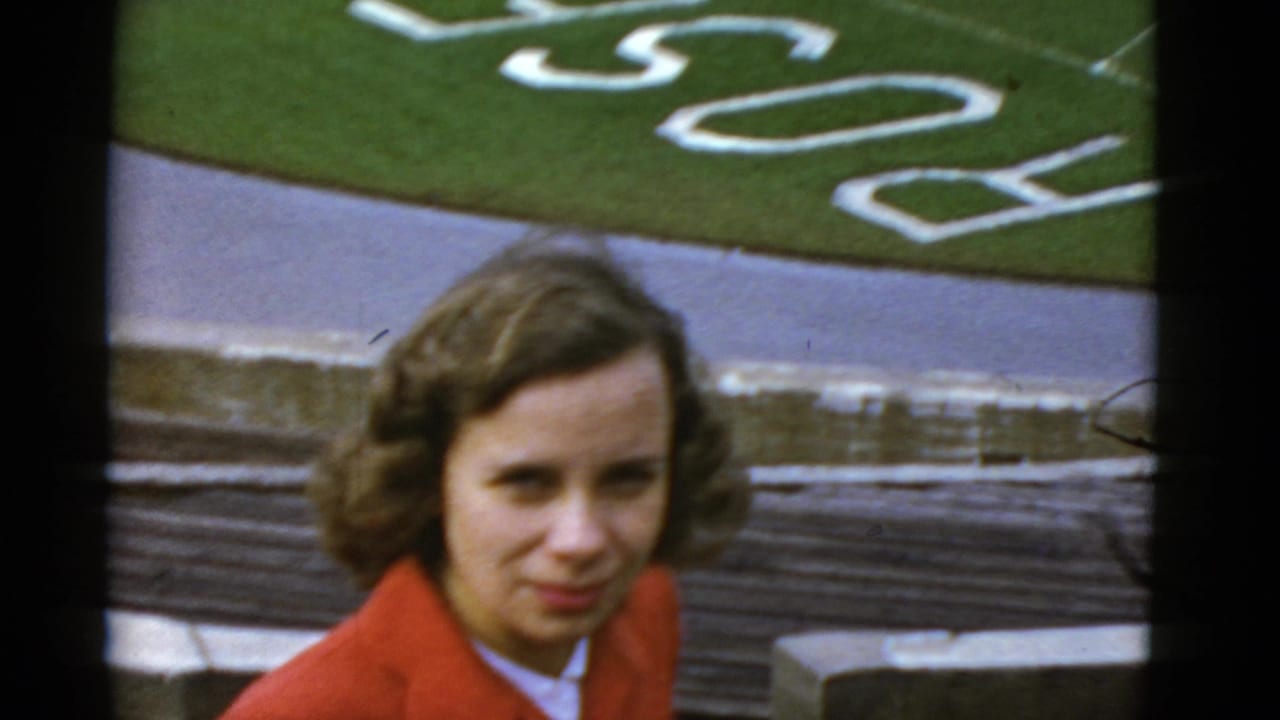 In a 1952 California home-movie frame, a young woman in an orange coat stands near stadium seating and a football field marked PACIFIC.