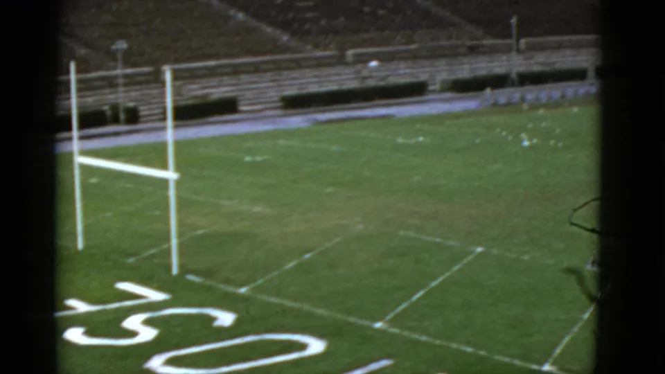 The camera looks across an empty California football field with goal posts, white yard lines, painted lettering, and stadium seating.