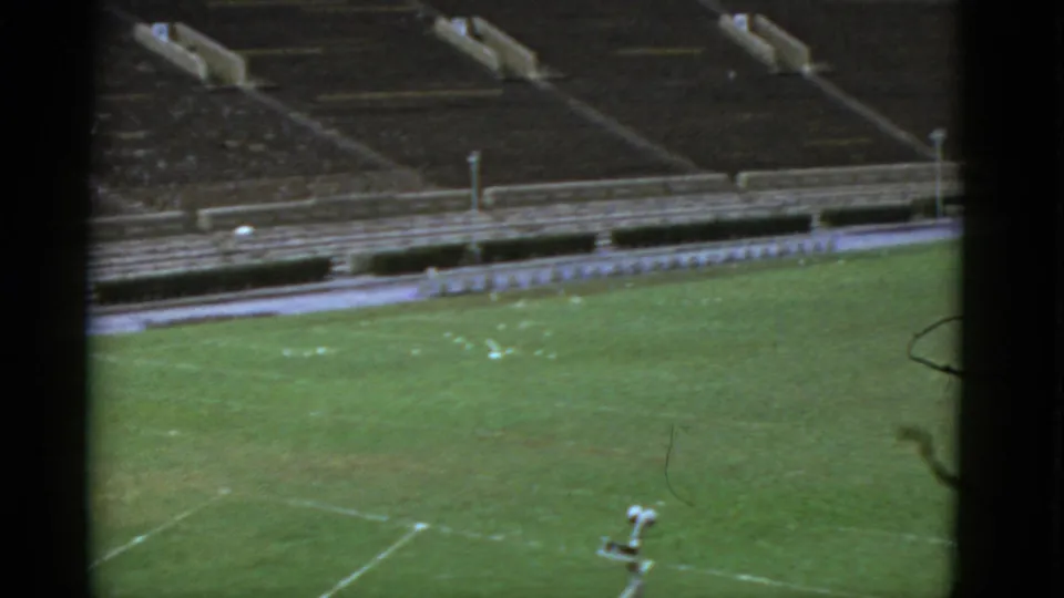 1952 California home-movie frame of a quiet football field from an elevated viewpoint, with pale markings and empty bleacher rows.