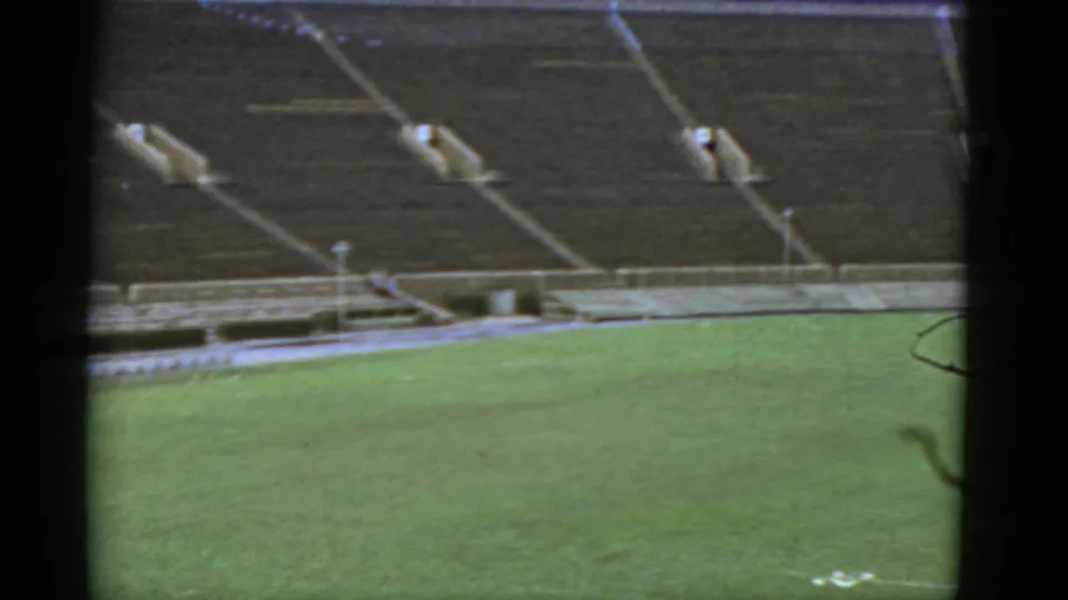 Archival 8mm frame from 1952 California showing a quiet stadium bowl, green field, and empty stepped seating.