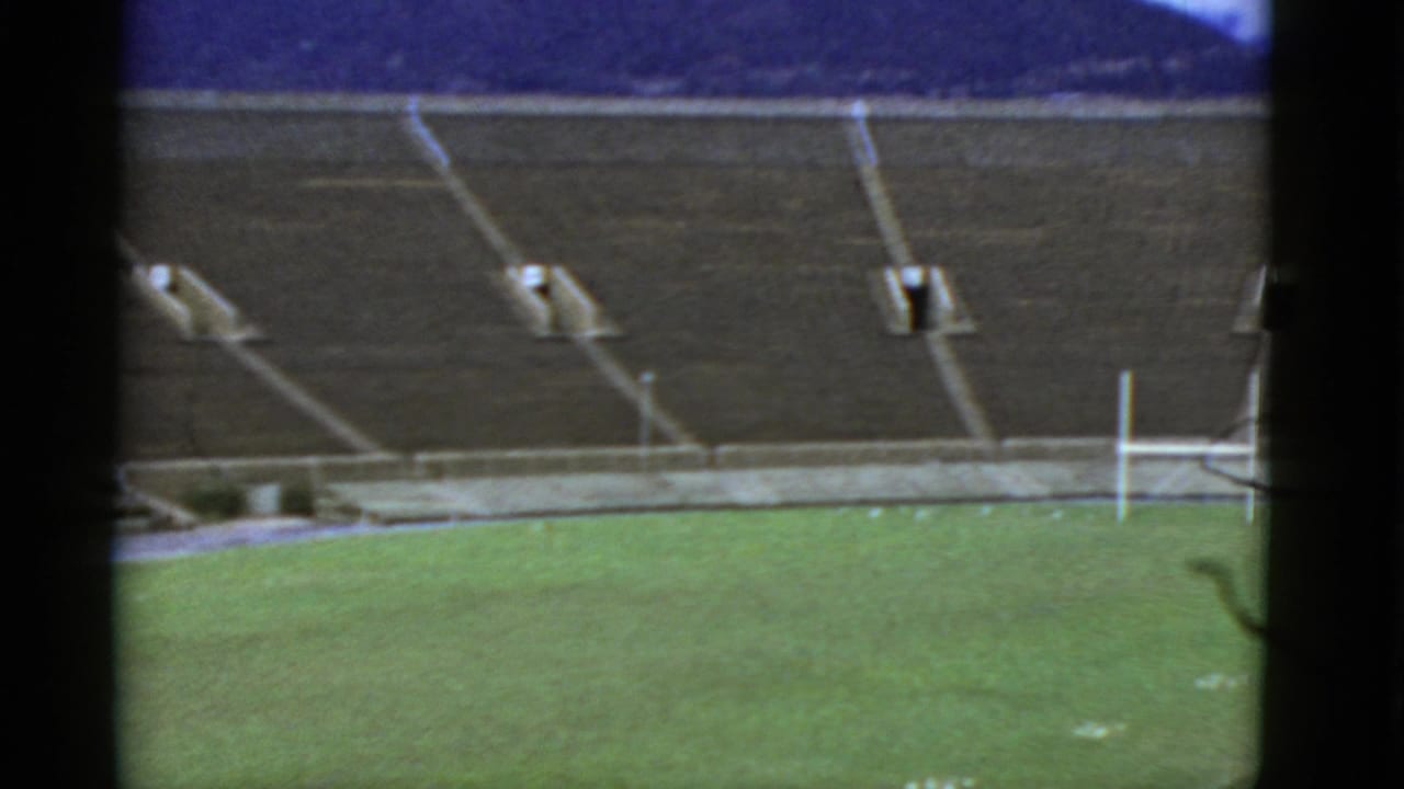 Empty stadium seating above a green field with white goalposts and hills beyond.