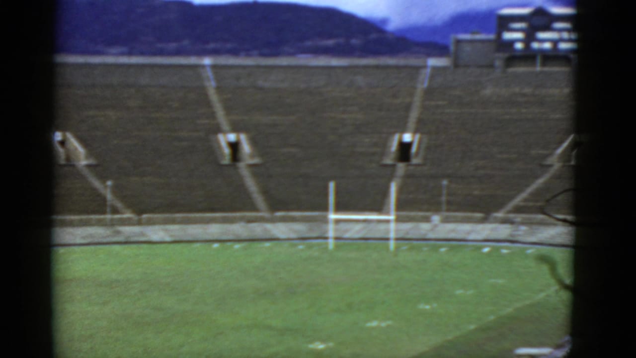 1952 home-movie frame of an empty stadium in California, with green turf, white goalposts, concrete stands, and a partial structure beyond.