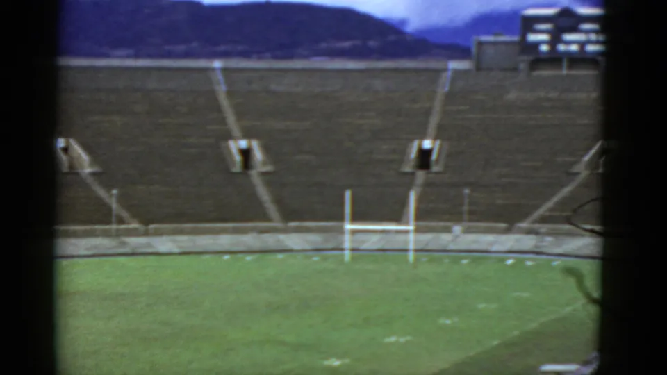 1952 home-movie frame of an empty stadium in California, with green turf, white goalposts, concrete stands, and a partial structure beyond.