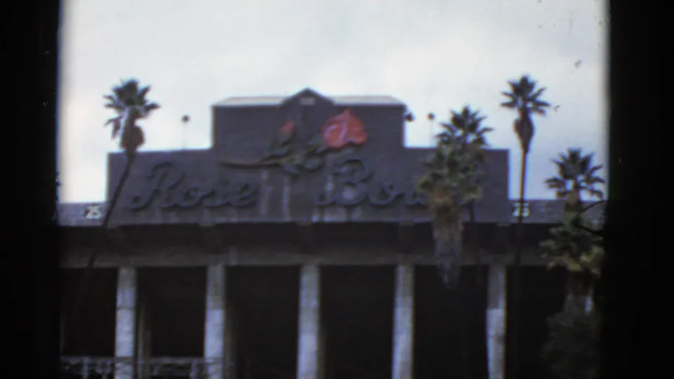 Static 1952 California home-movie frame of the Rose Bowl facade, cursive lettering, red rose emblem, palms, and dark foreground bars.