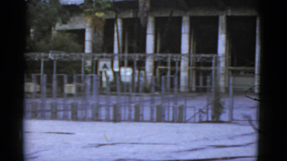 1952 California home-movie frame showing a metal gate, fenced enclosure, trees, and a columned public-venue building.