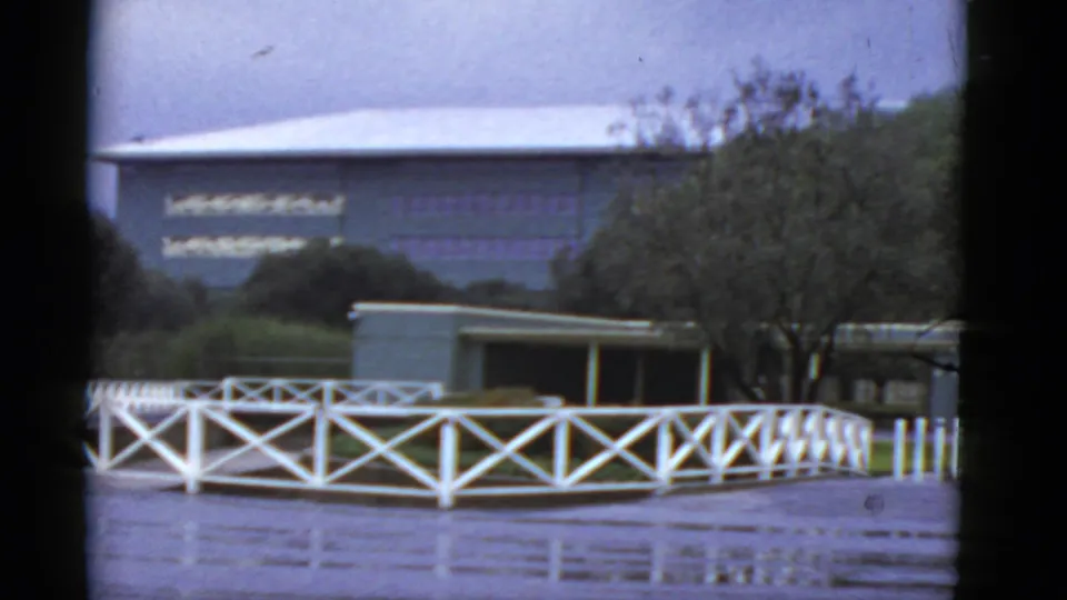 1952 California home-movie frame showing a white rail fence, trees, low venue structures, and overcast sky.