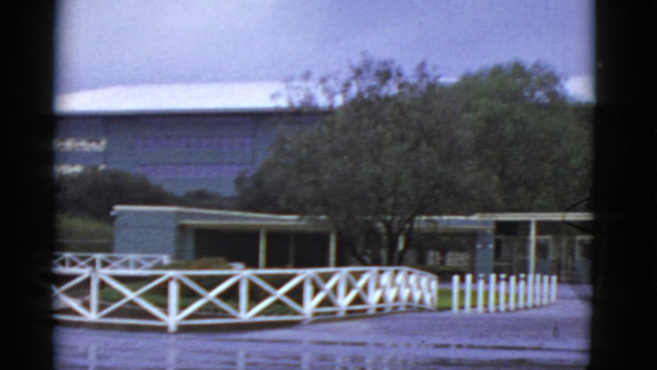 Flat-roof building behind a white X-pattern fence and trees.